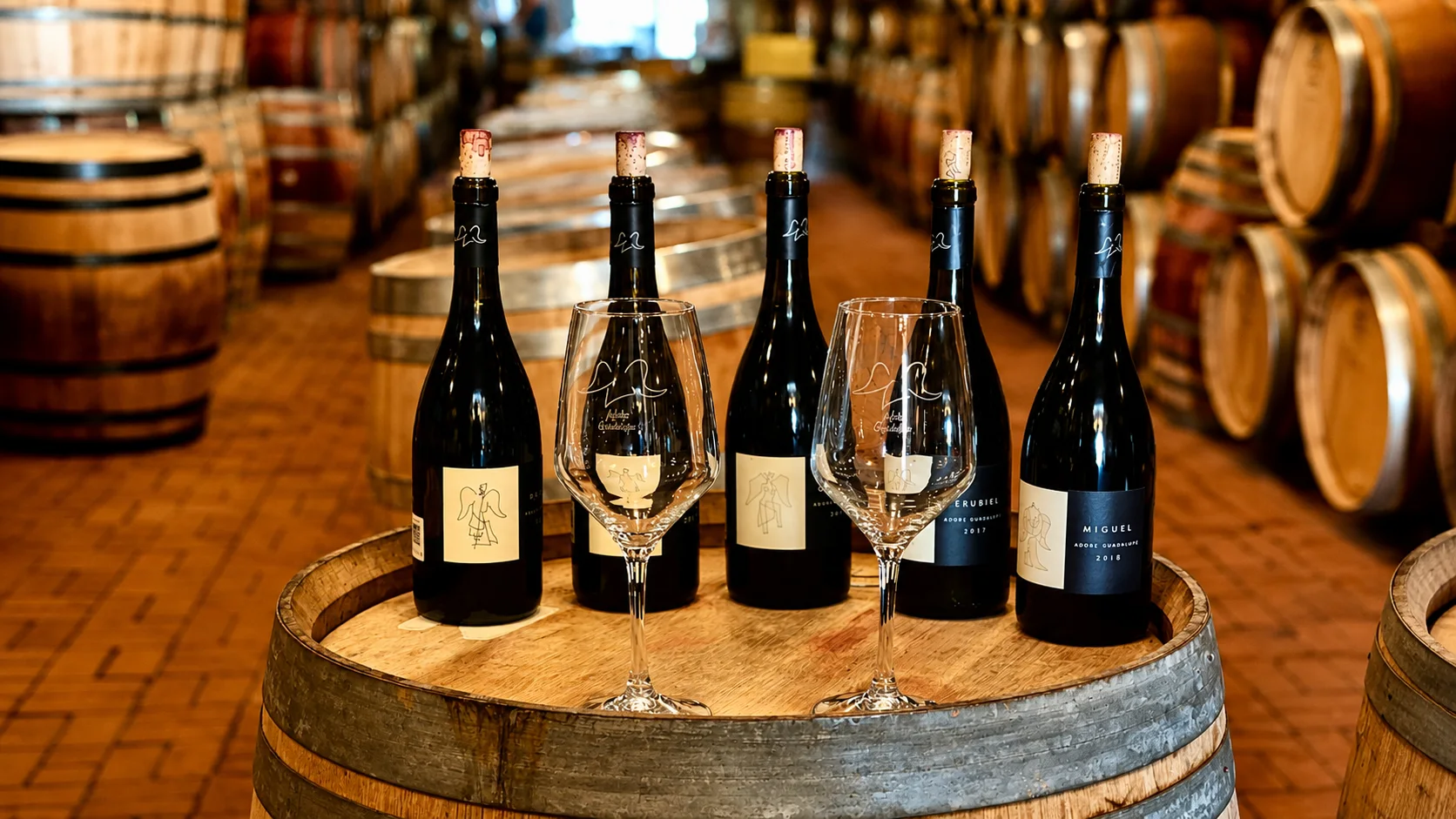 Luxury wine tours at Adobe Guadalupe with five wine bottles and branded glasses arranged on a cellar barrel.