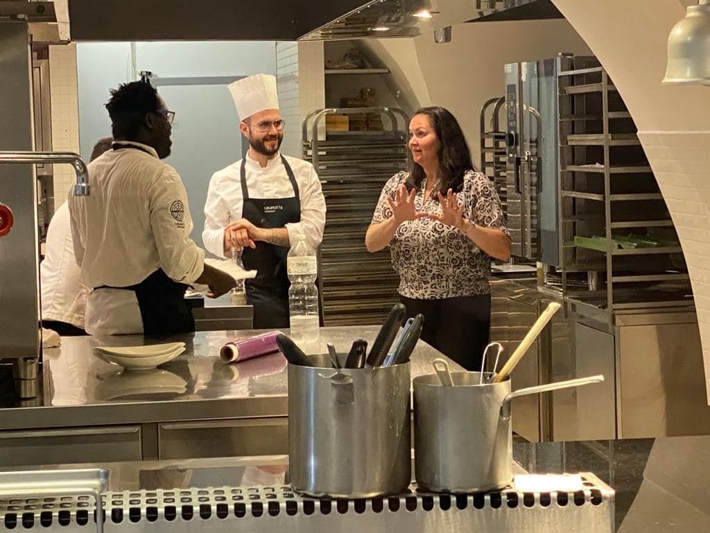 Two chefs and a woman talking in a professional Casamata kitchen with stainless steel counters and equipment.